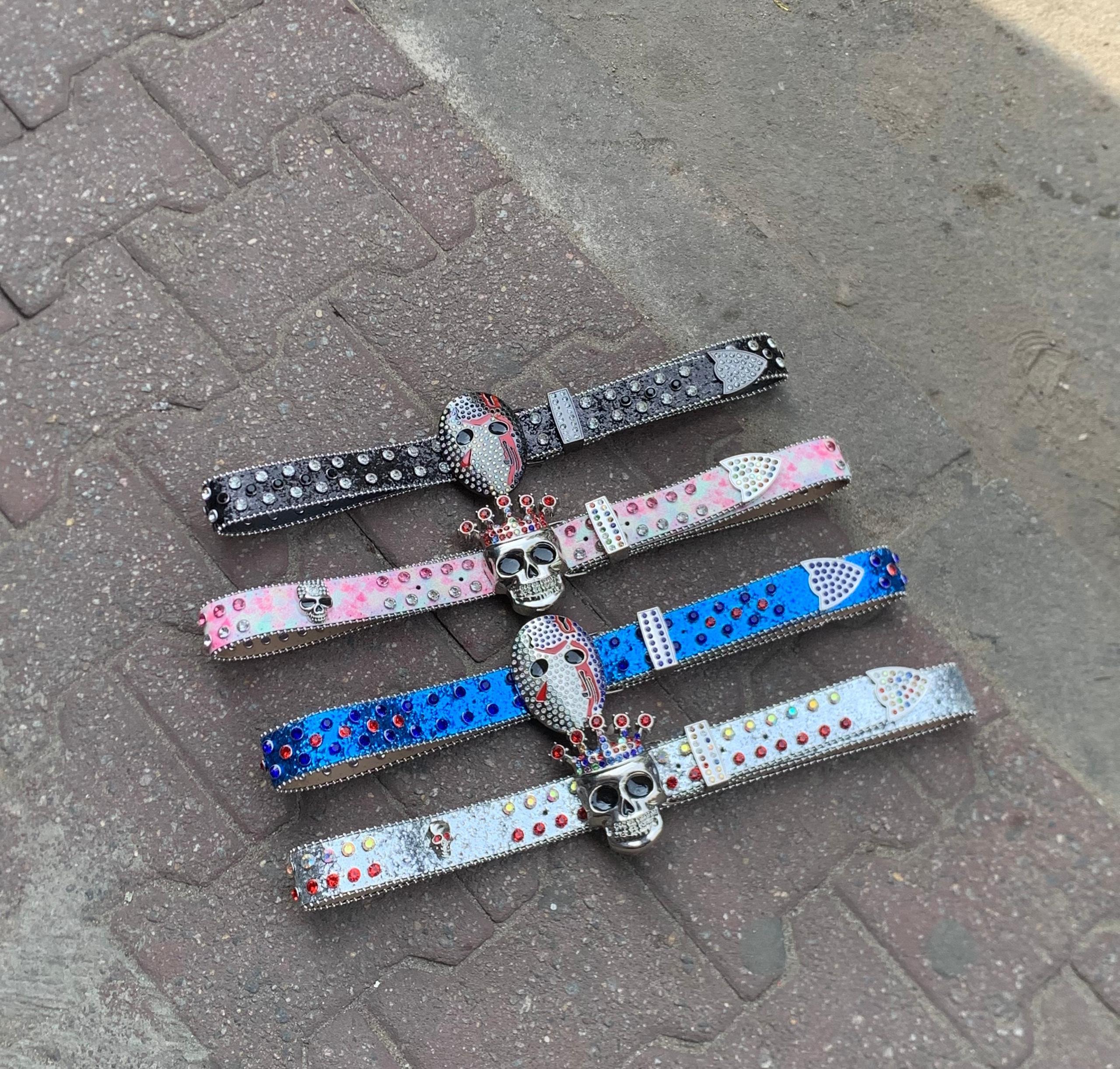 Four designer belts with rhinestones and skull or hockey mask buckles in black, pink, blue, and silver laid on pavement.