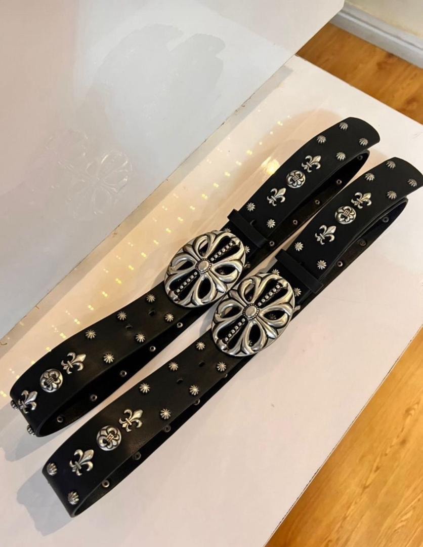 Black leather belts with large silver flower buckles, fleur-de-lis and star details on white surface.