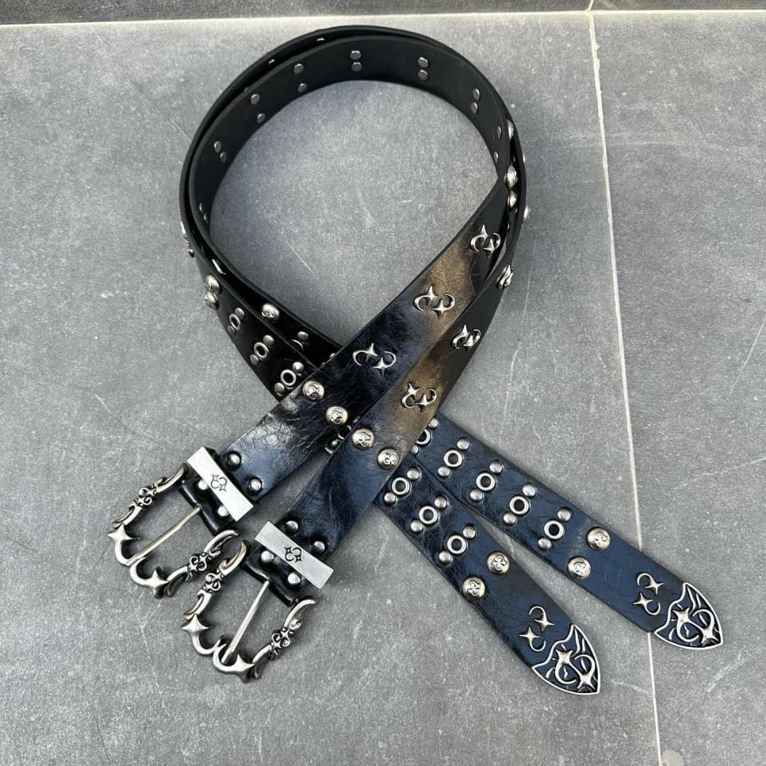 Black leather belt with ornate silver anchor buckles, studs and emblems arranged on gray surface.