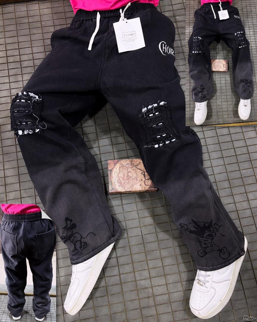 Model wearing black graphic ripped denim joggers with CHOR text and cartoon prints on legs.