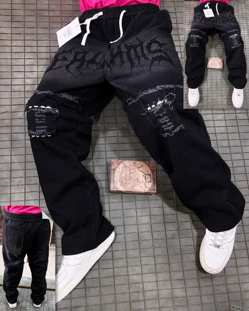 Model in black denim joggers with large gothic print on thigh and ripped knee patches, styled with white sneakers.