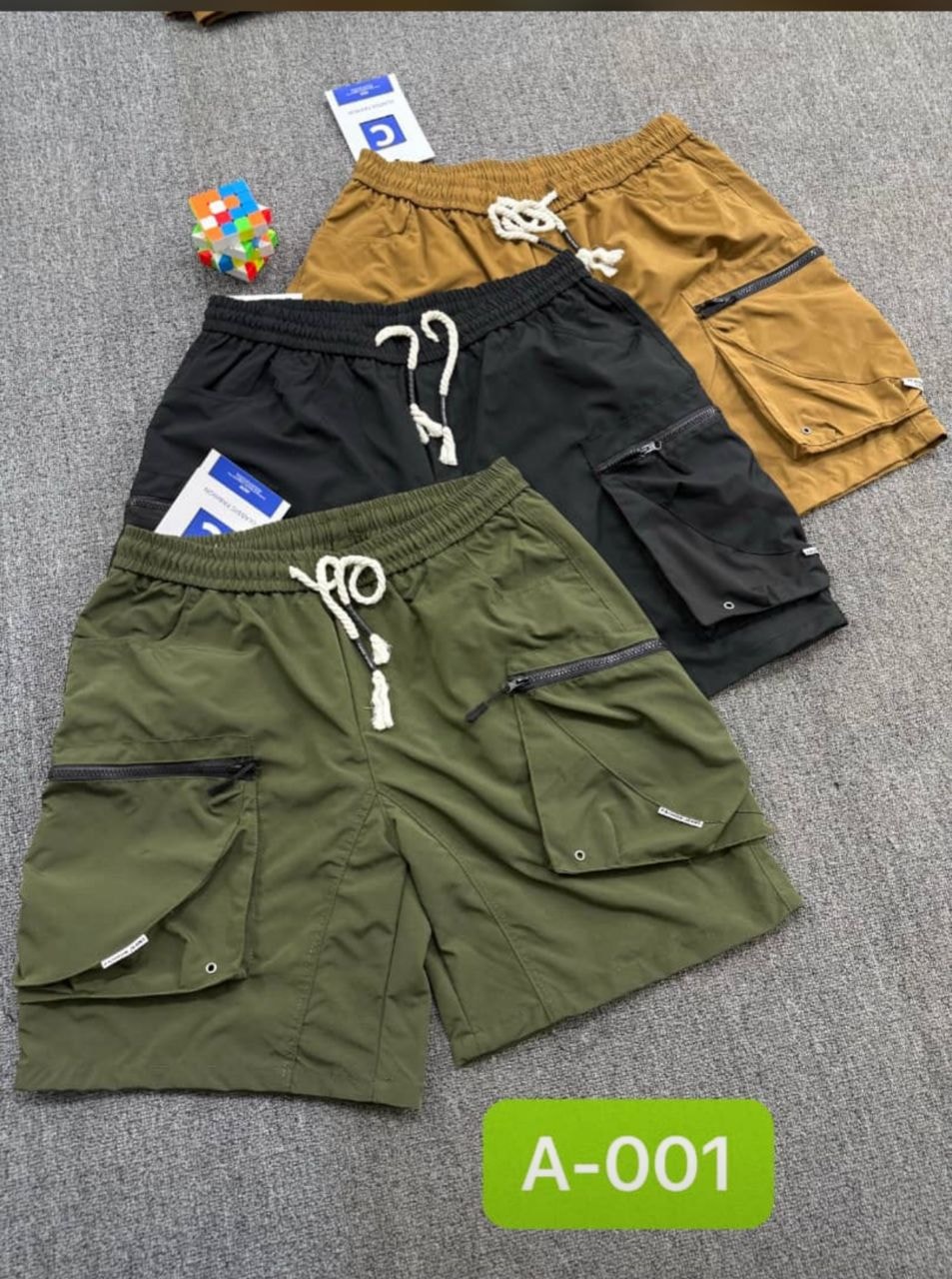 Three cargo combat shorts in olive, black and khaki with zip pockets and drawstring waist laid flat.