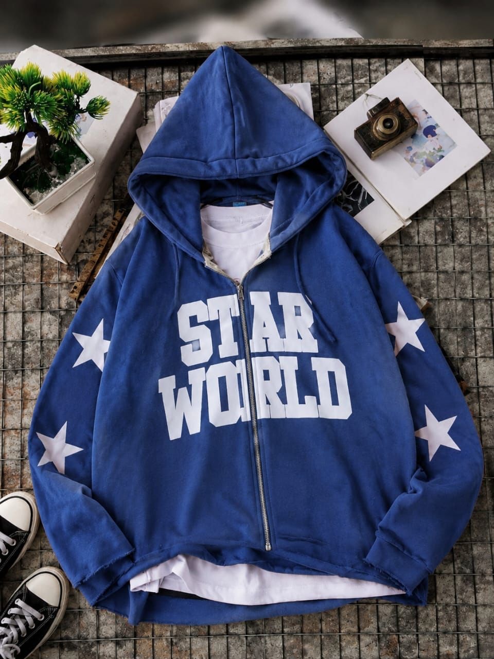 Blue zip-up hoodie with STAR WORLD text and white stars on sleeves in flat lay.