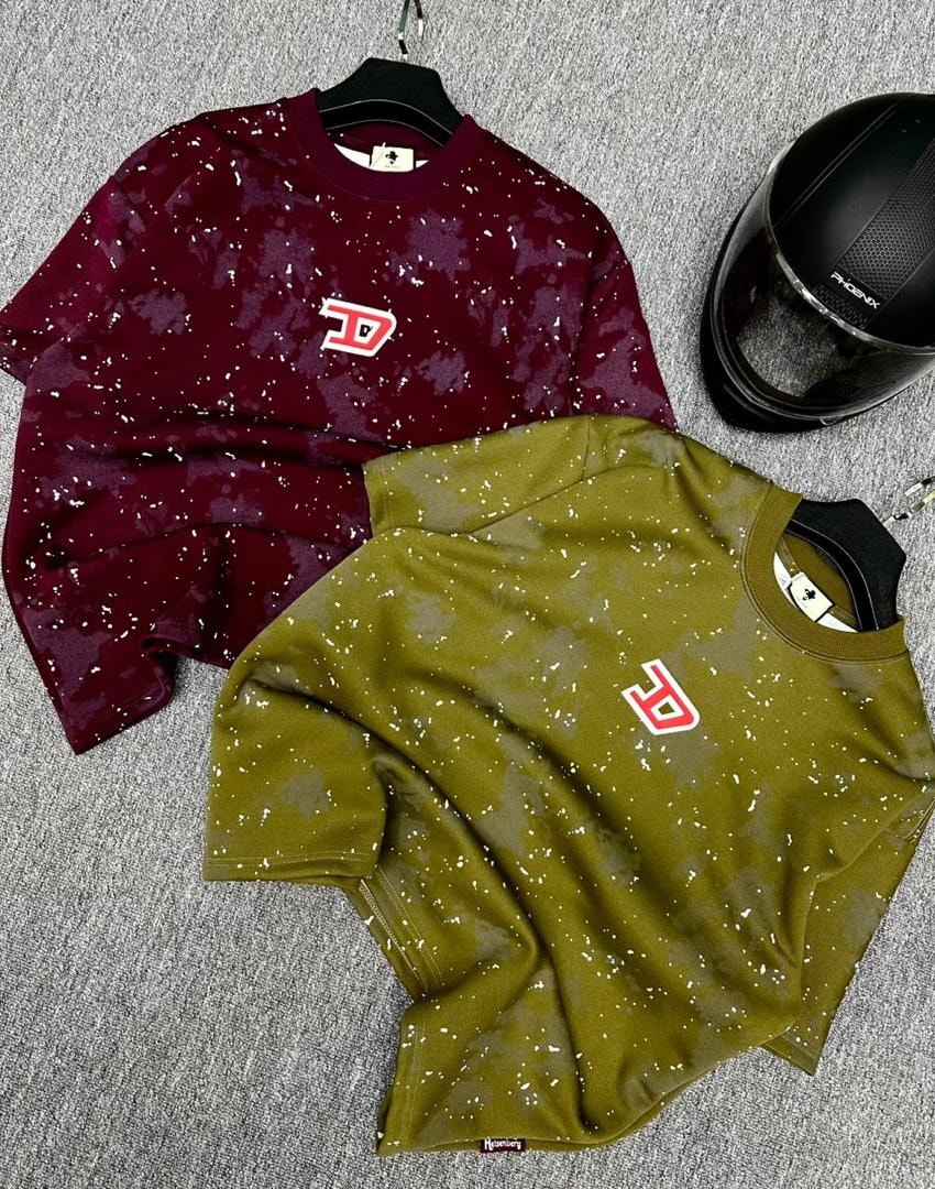 Maroon and olive splatter t-shirts featuring red D logo and tags beside a motorcycle helmet.