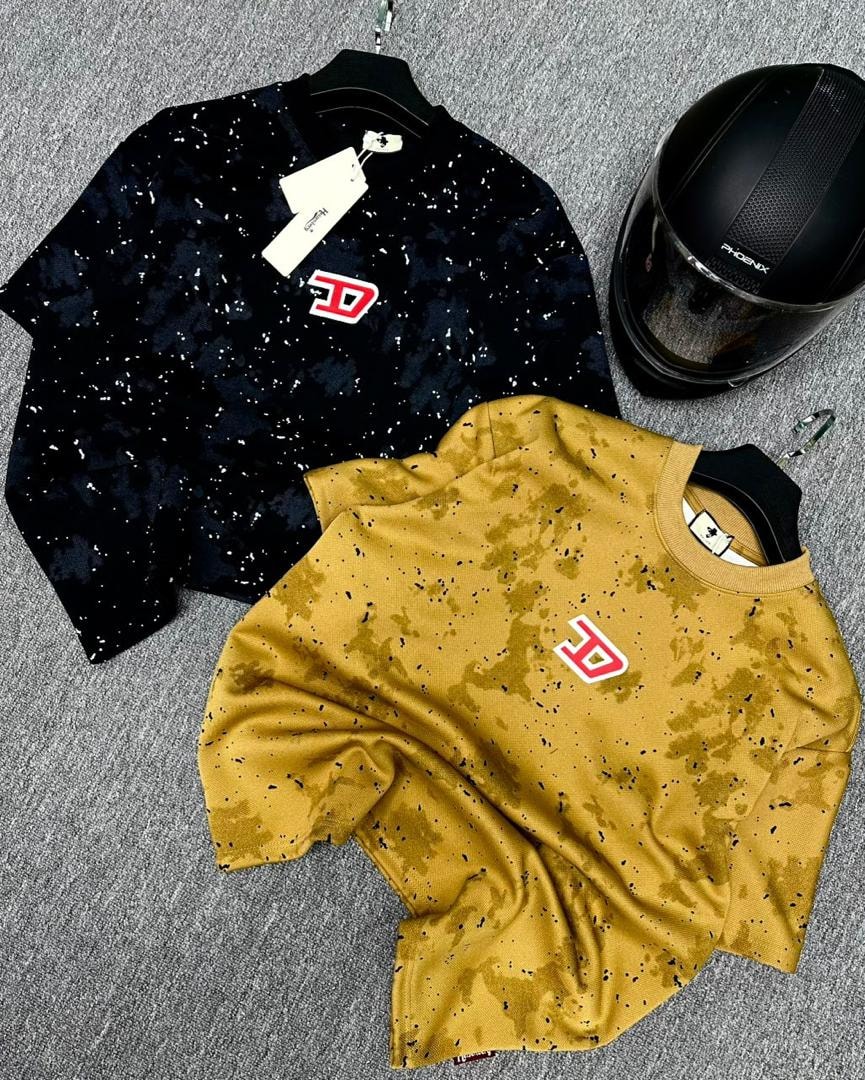 Black and mustard splatter t-shirts with red D logo displayed with black helmet on gray surface.