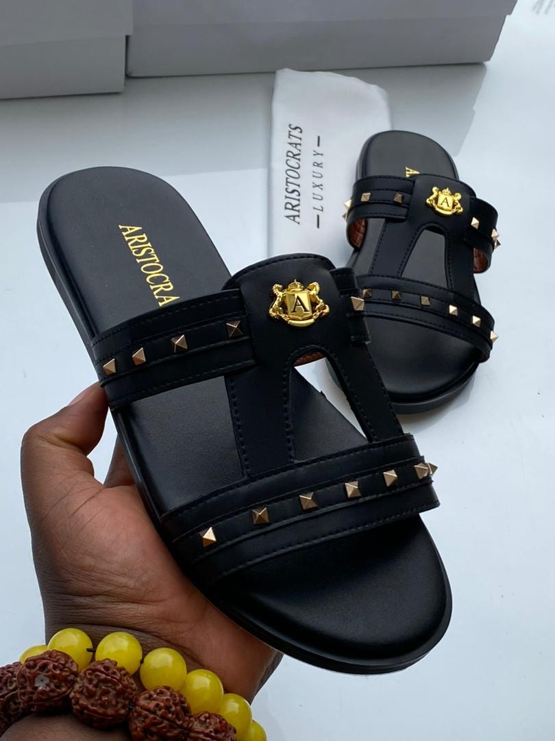 Hand holding black Aristocrats studded slides showing gold emblem and multiple straps.