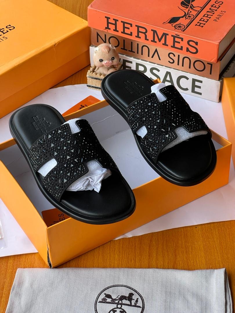 Black perforated Hermès slides displayed in orange luxury box with dust bag and accessories.