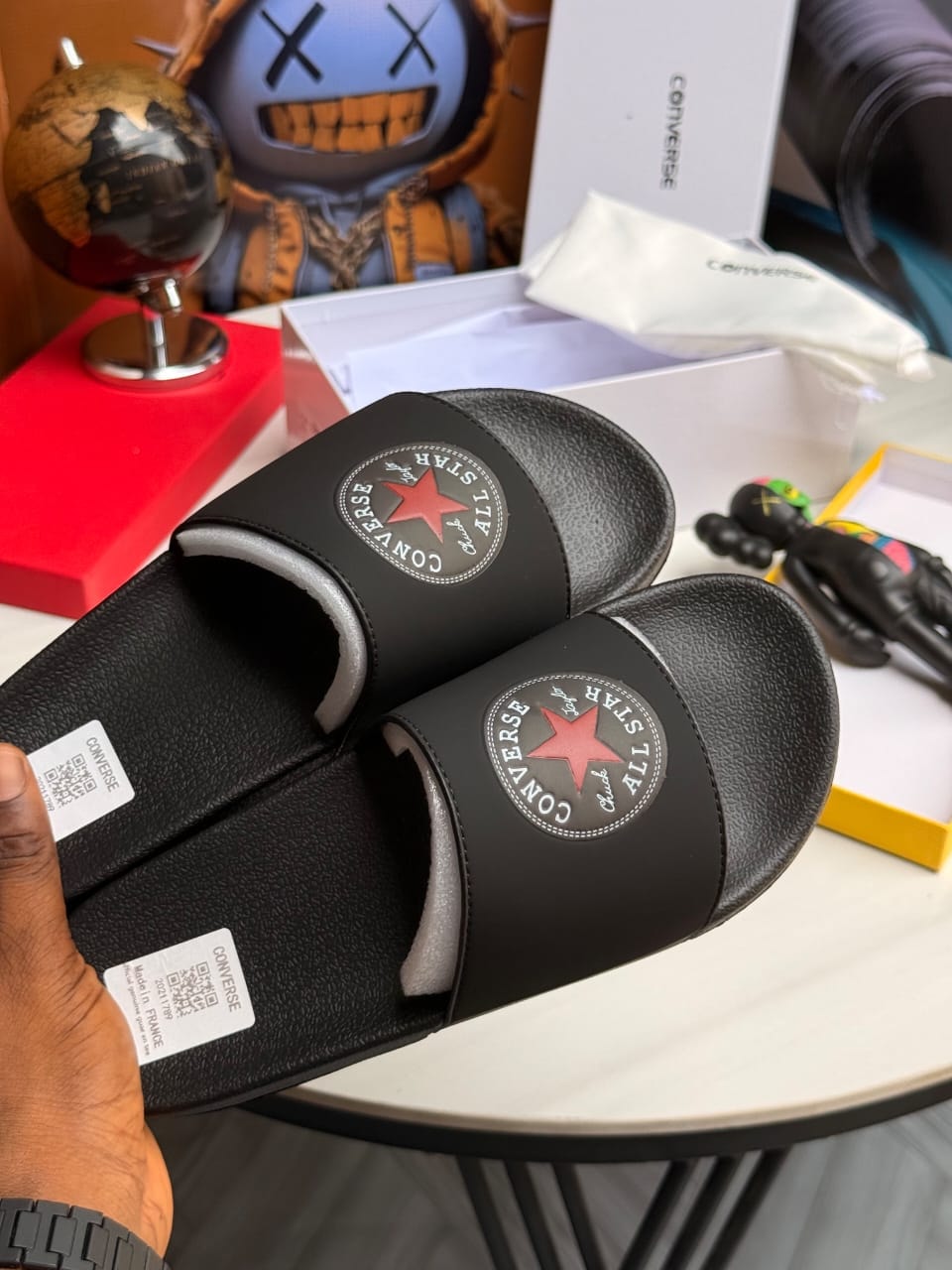 Hand holding pair of black Converse All Star slides with circular logo.