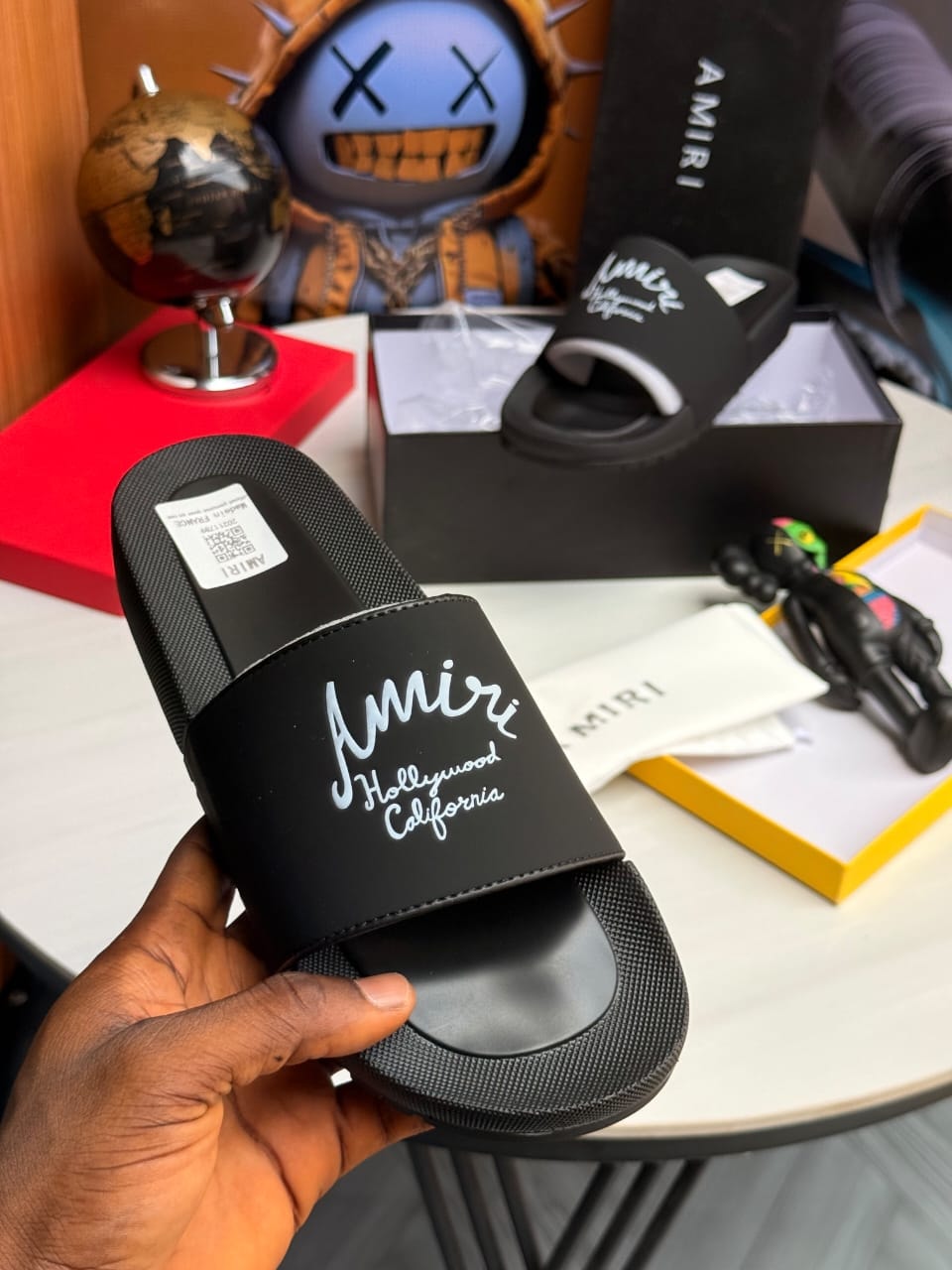 Hand holding black Amiri slide with white script text on strap.