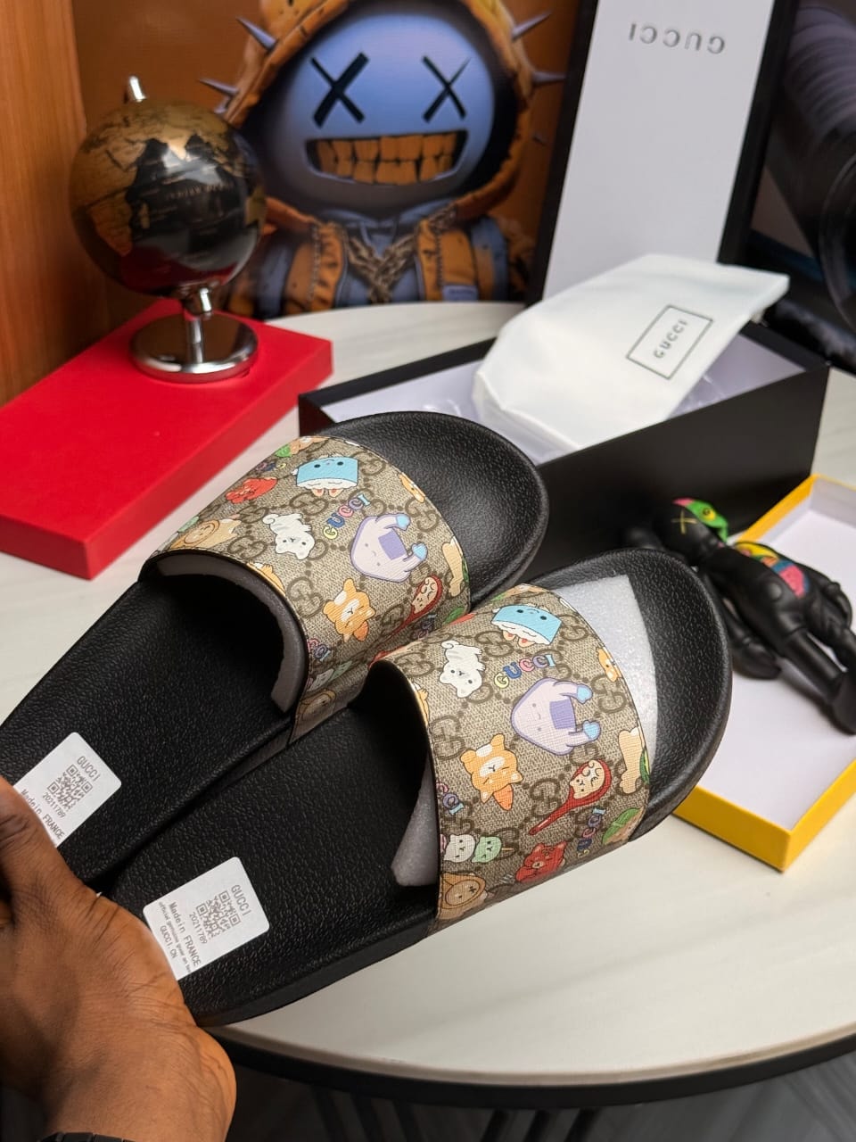 Hand holding beige Gucci slide with colorful cartoon prints and monogram.