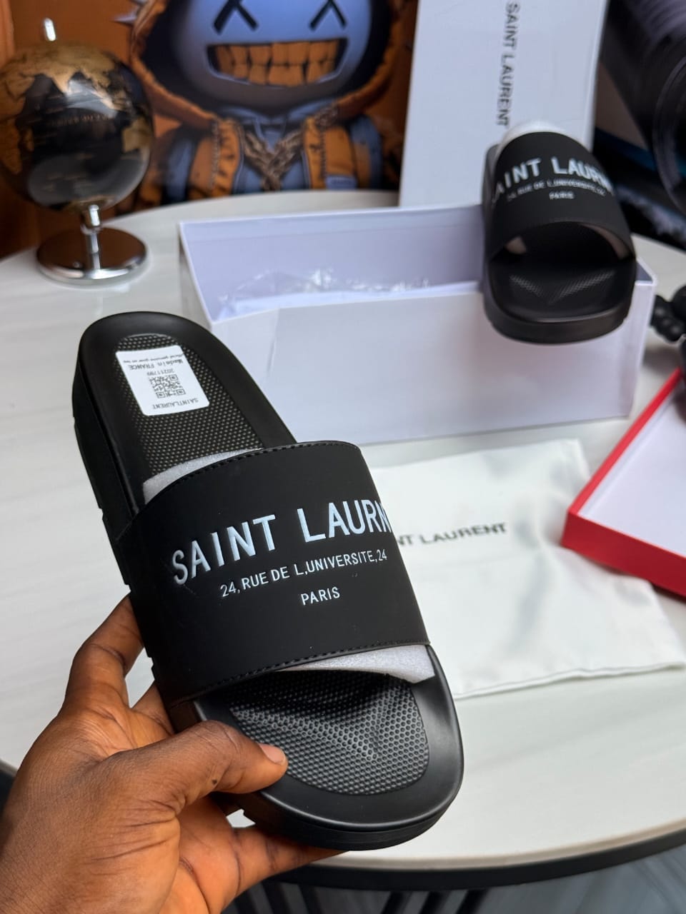 Hand holding black Saint Laurent slide with white text branding.