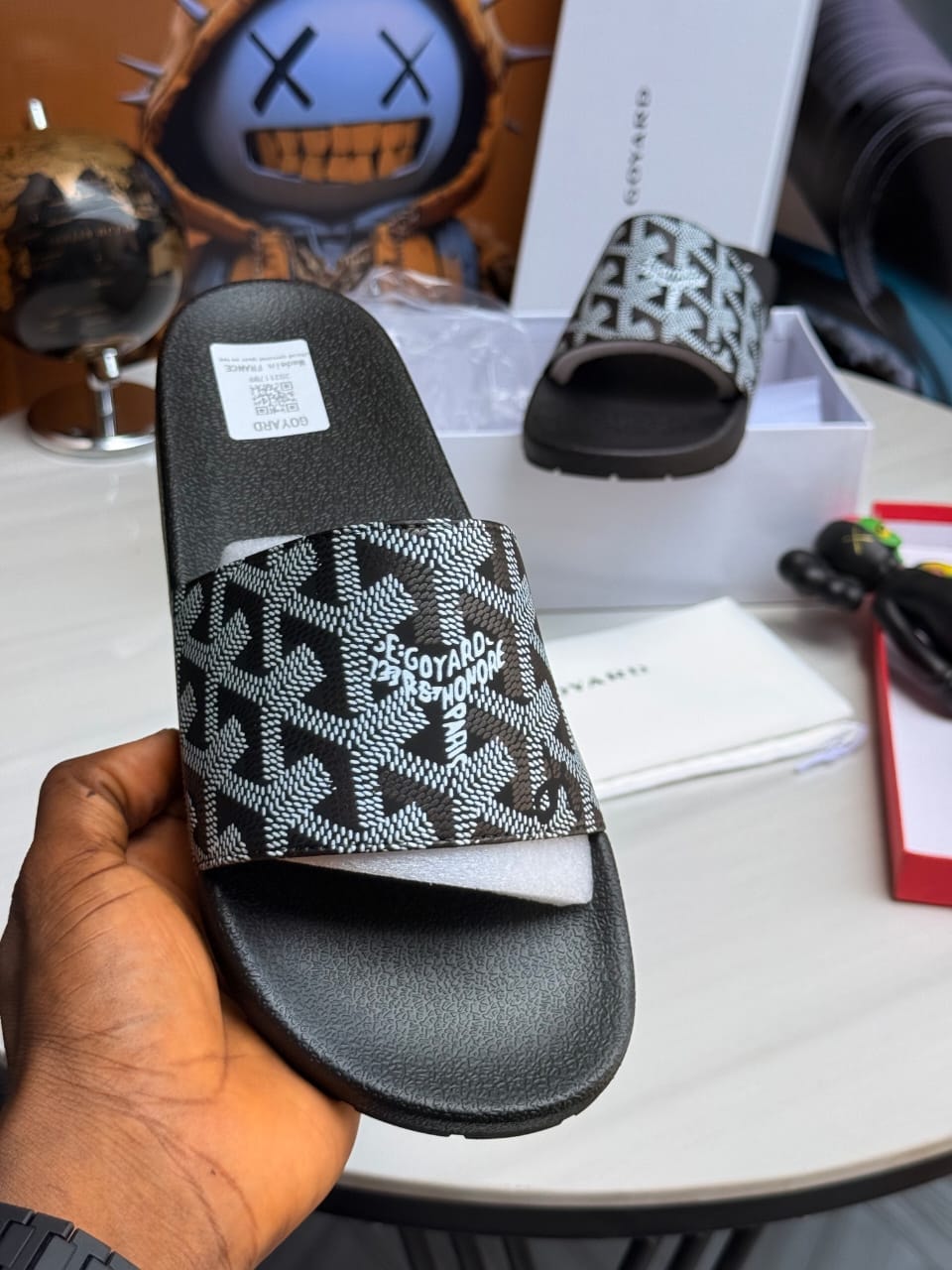Hand holding black and white patterned Goyard slide with text and tag.