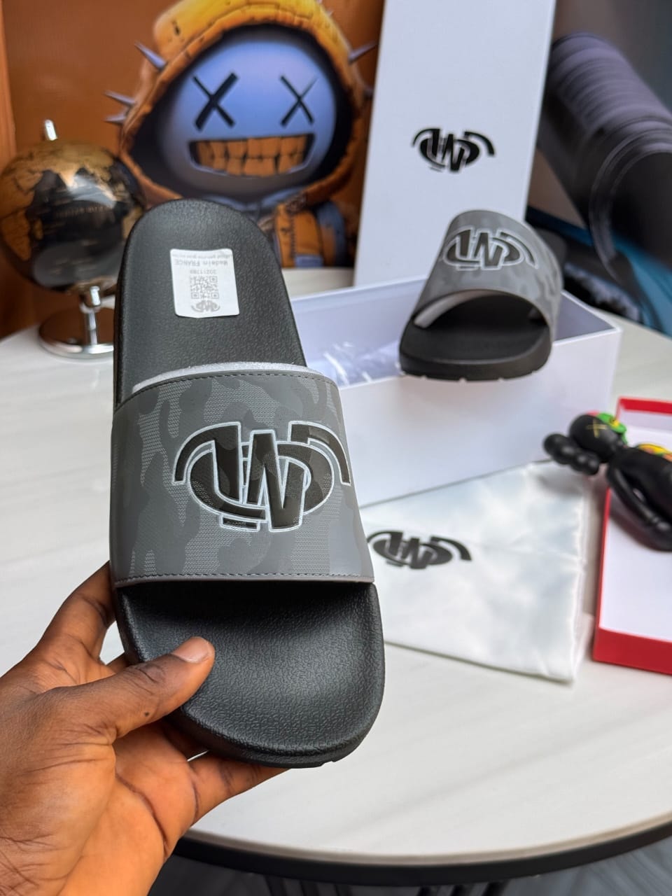 Hand holding gray camouflage slide with large logo on strap and visible tag.