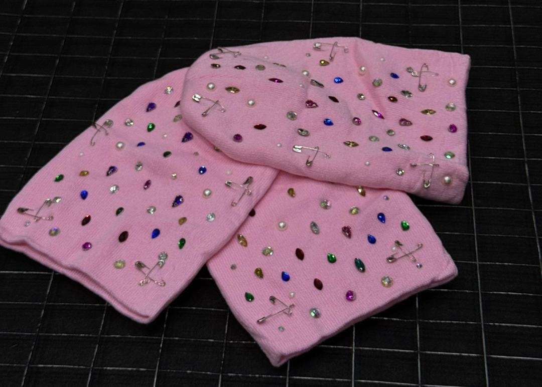 Two pink rhinestone embellished beanies with colorful stones and safety pins on black grid surface.