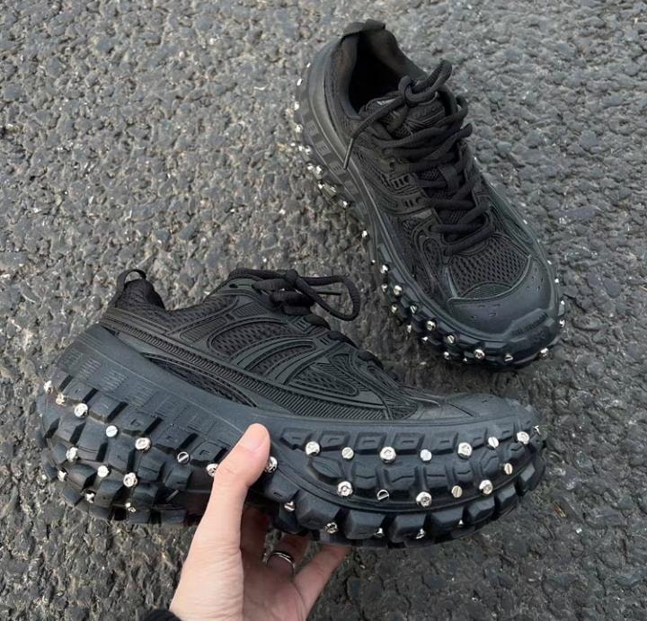 Black studded sneakers held by hand showing sole details on asphalt.