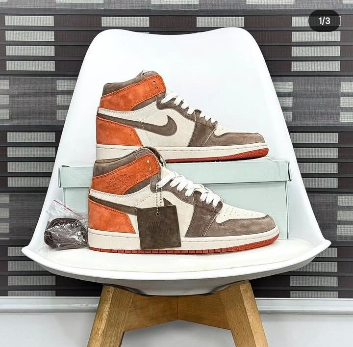 Nike Air Jordan 1 high-top sneakers in white, brown and orange color blocking displayed on a white chair with box and tags.