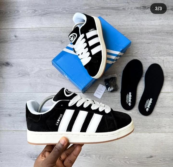 Black Adidas Campus sneakers with white stripes shown with blue shoebox, extra insoles, and accessories on wooden surface.