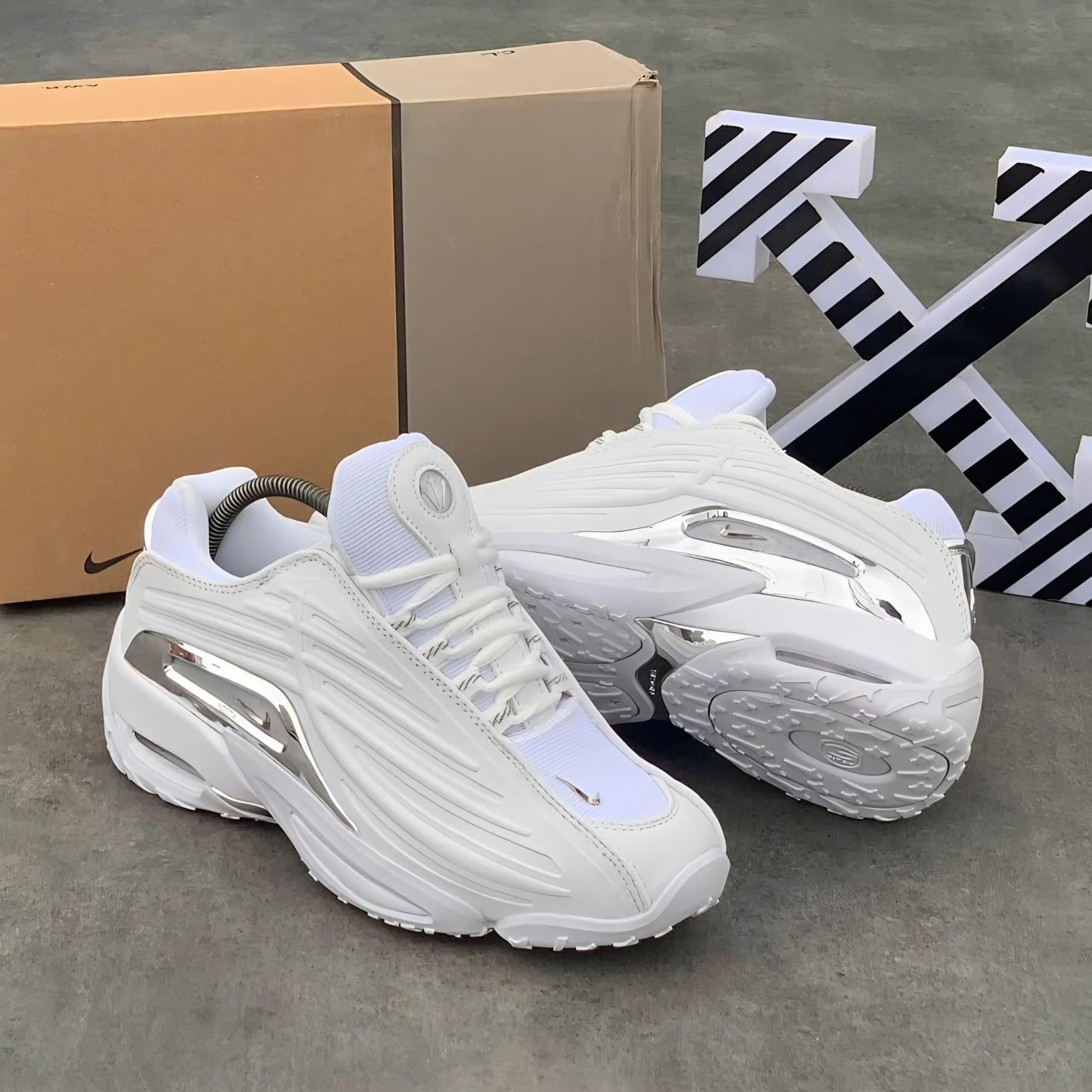 White and chrome Nike NOCTA Hot Step sneakers displayed with original box and decorative arrows.