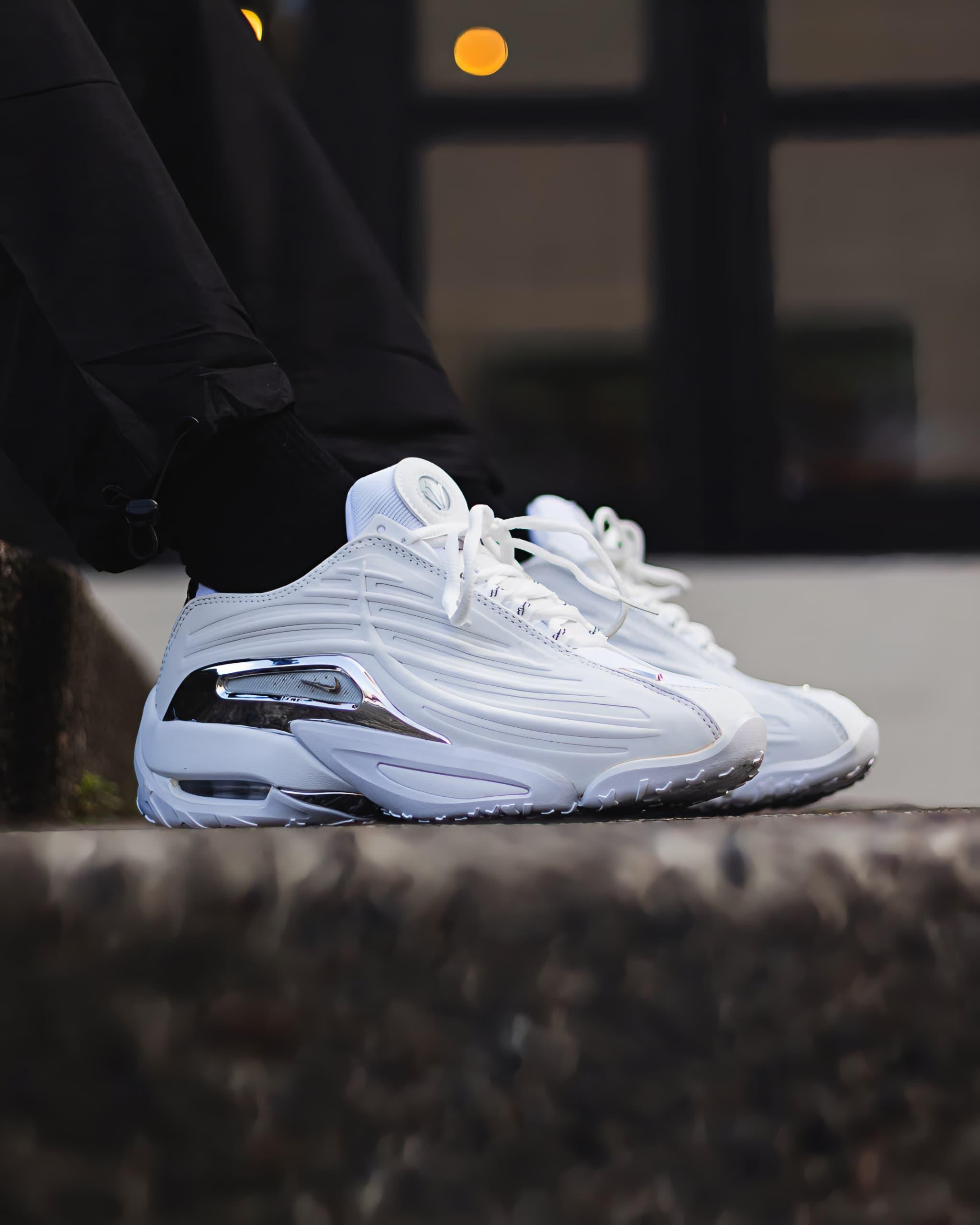 Person wearing white Nike NOCTA Hot Step sneakers with chrome accents outdoors.