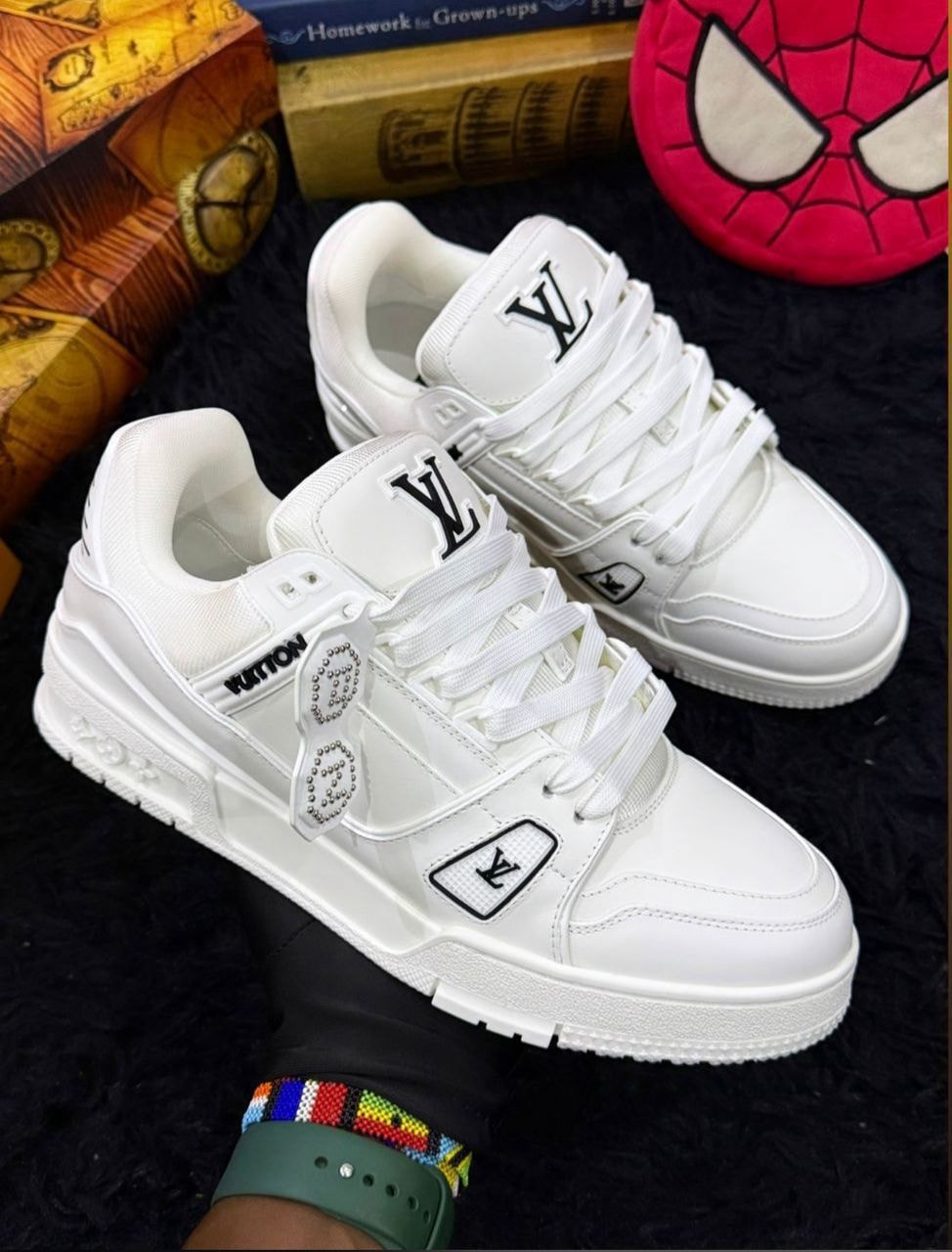 Pair of white Louis Vuitton sneakers held on dark surface with books and decor in background.