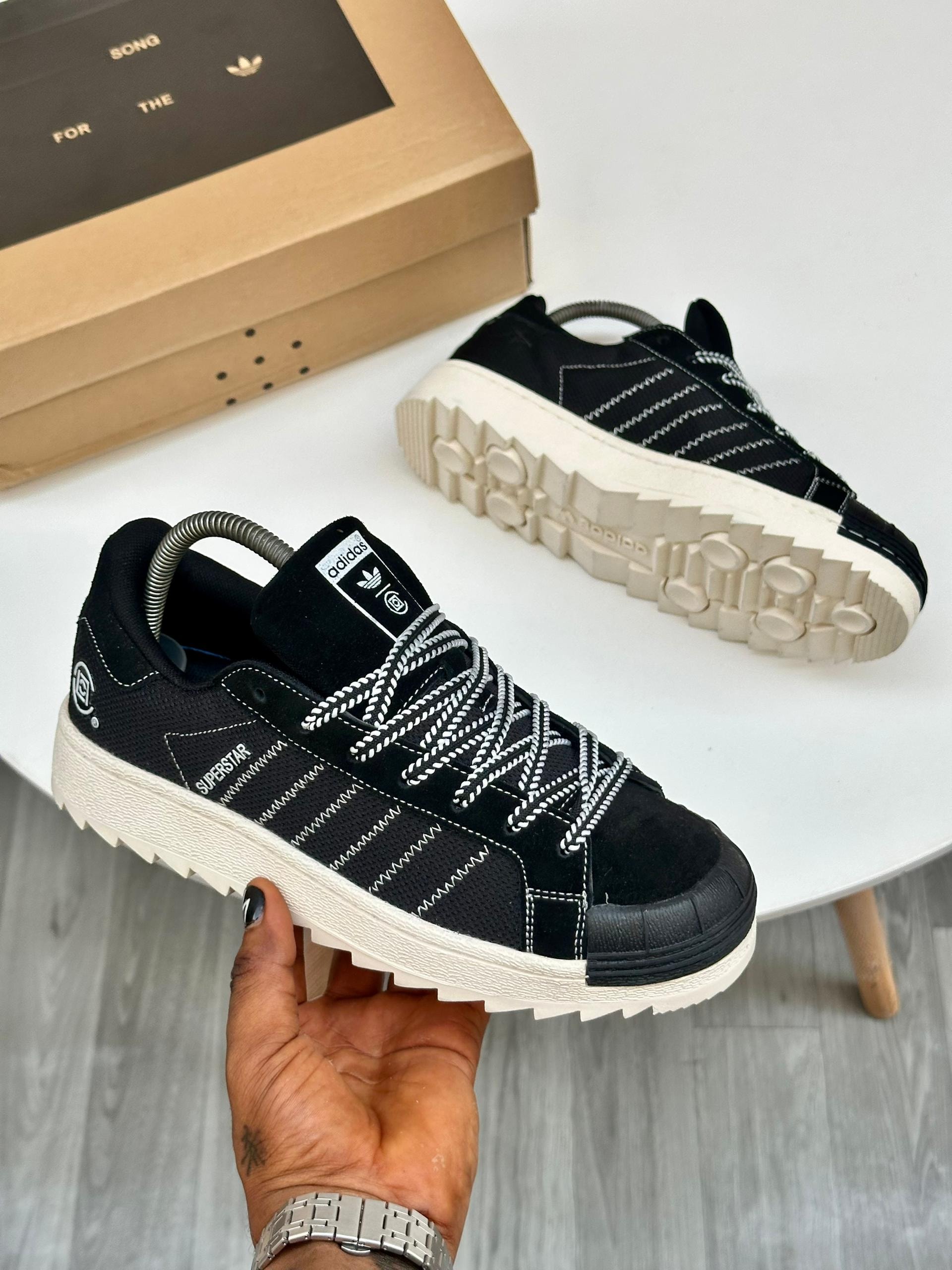 Black Adidas Superstar sneakers with white stitching and chunky sole held in hand with branded box.