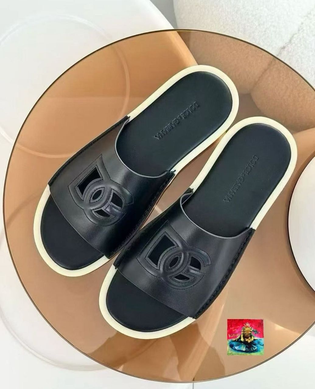 Black Dolce & Gabbana slides with large logo on strap and white sole on glass table.