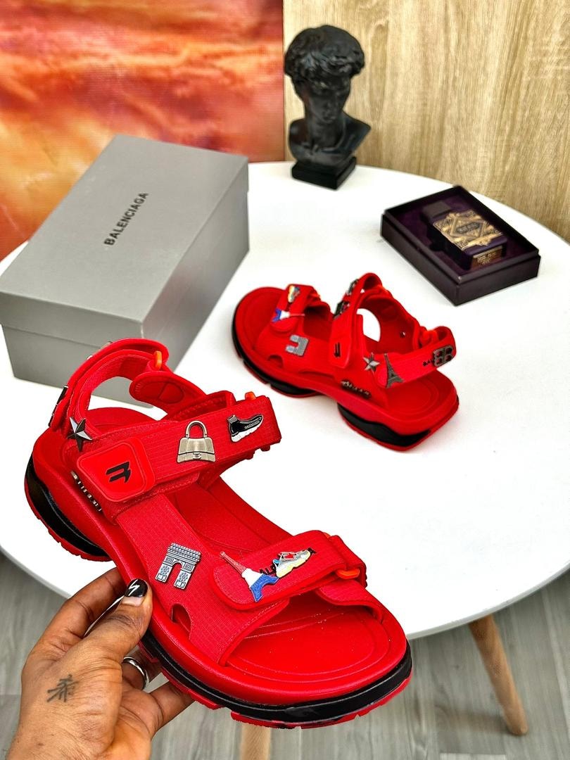 Red Balenciaga style sandals with multiple straps, charms and decorative elements held in hand with branded box.