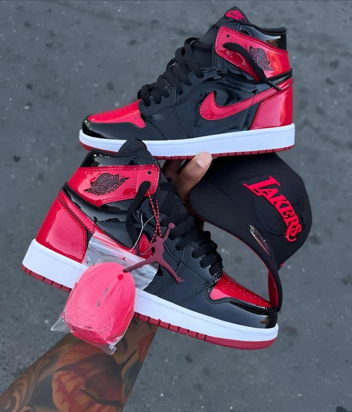 Red and black patent Nike Air Jordan 1 High sneakers held with red laces, Jumpman charm and Lakers cap.