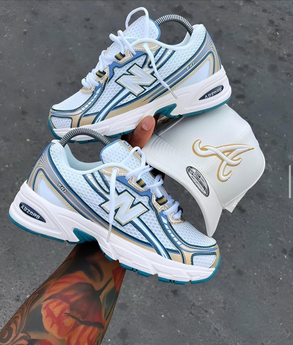 New Balance 1000 sneakers held in hand showing side profile with white and gold cap accessory.