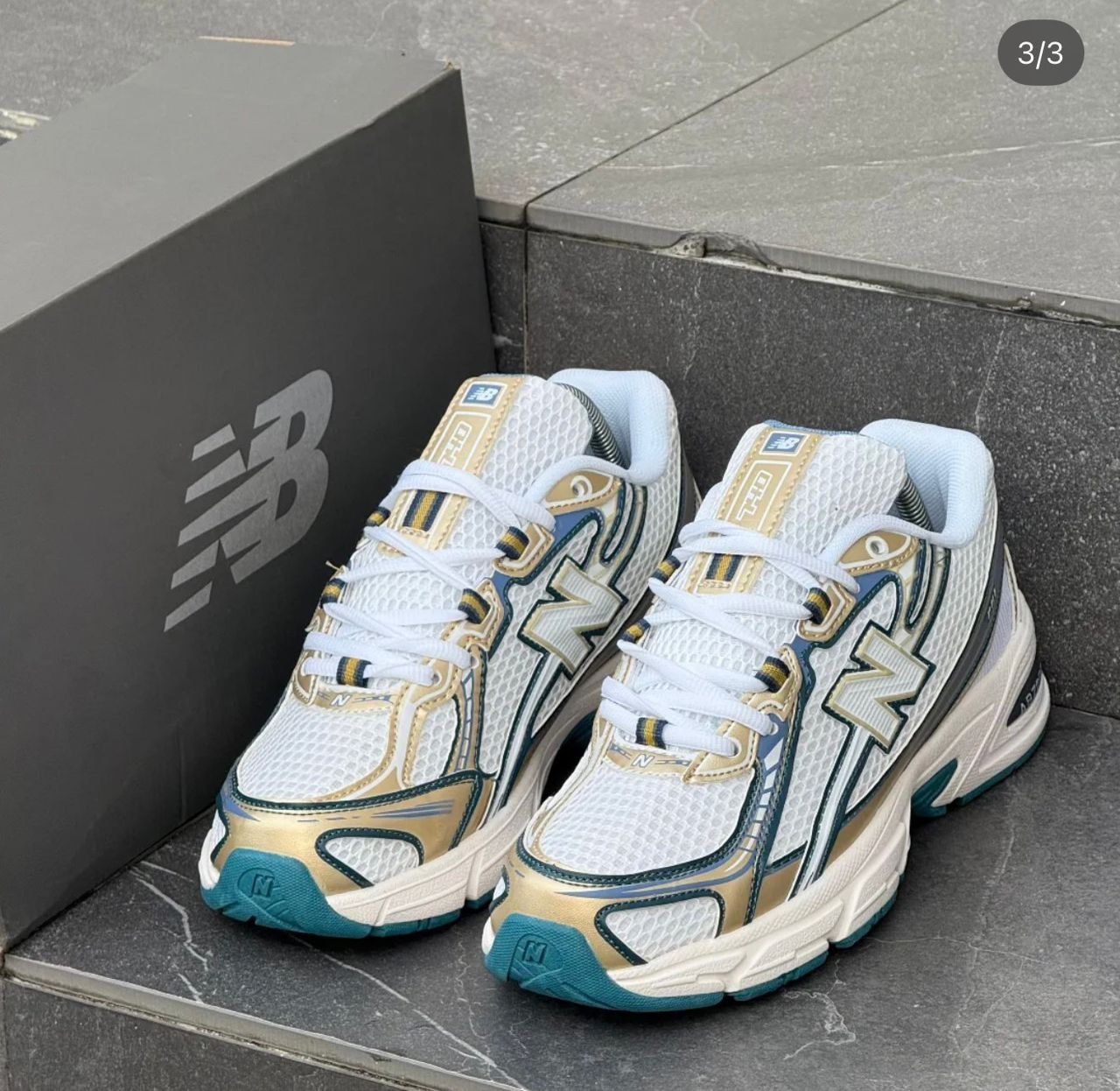 New Balance 1000 sneakers in white, gold and teal on steps beside branded box.
