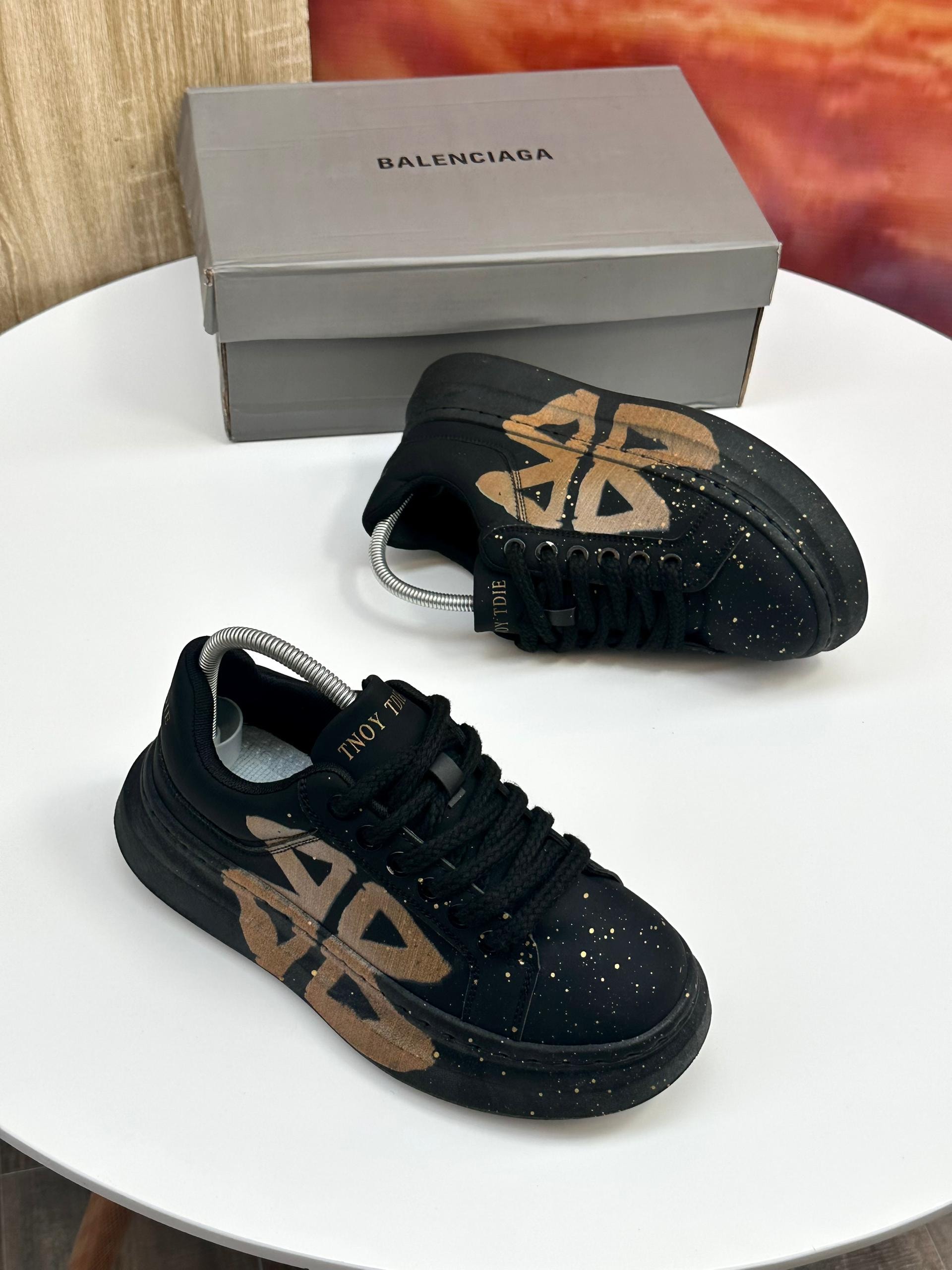 Black TNOY TDIE sneakers with gold graphics and speckles on white table with Balenciaga box.