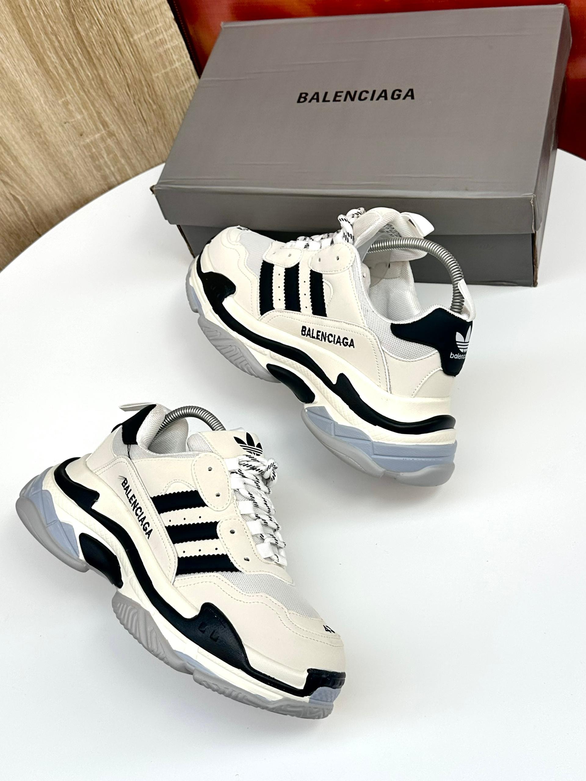 White black Balenciaga Adidas chunky sneakers on white table with grey branded box in background.