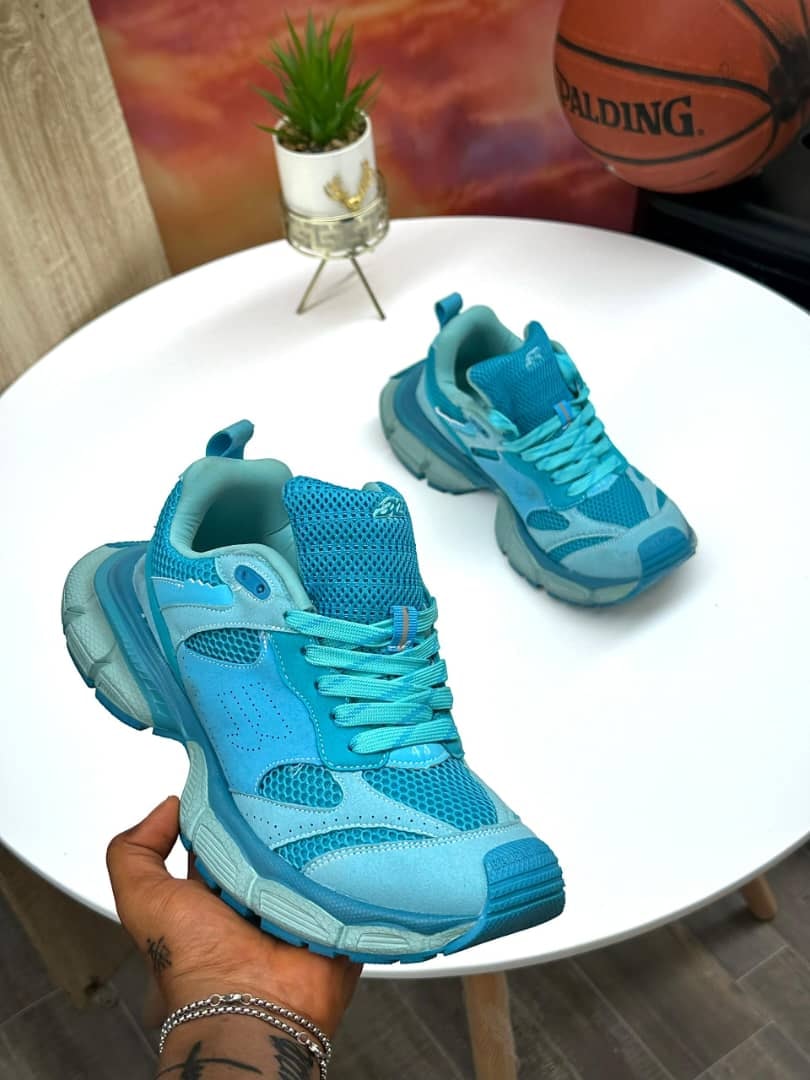 Bright blue chunky sneakers held in hand and shown on white table with plant and basketball in background.