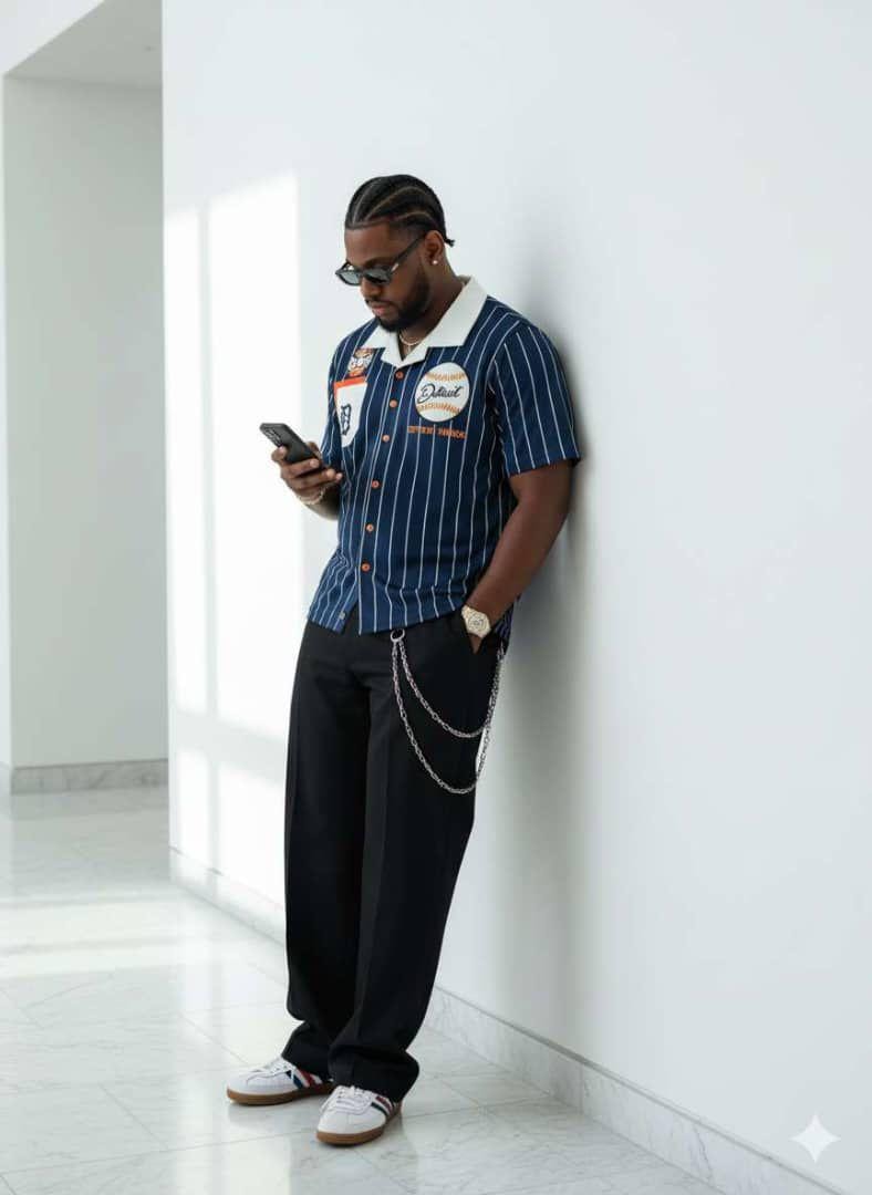 Man modeling blue striped baseball shirt with patches, paired with black trousers and chain accessory.