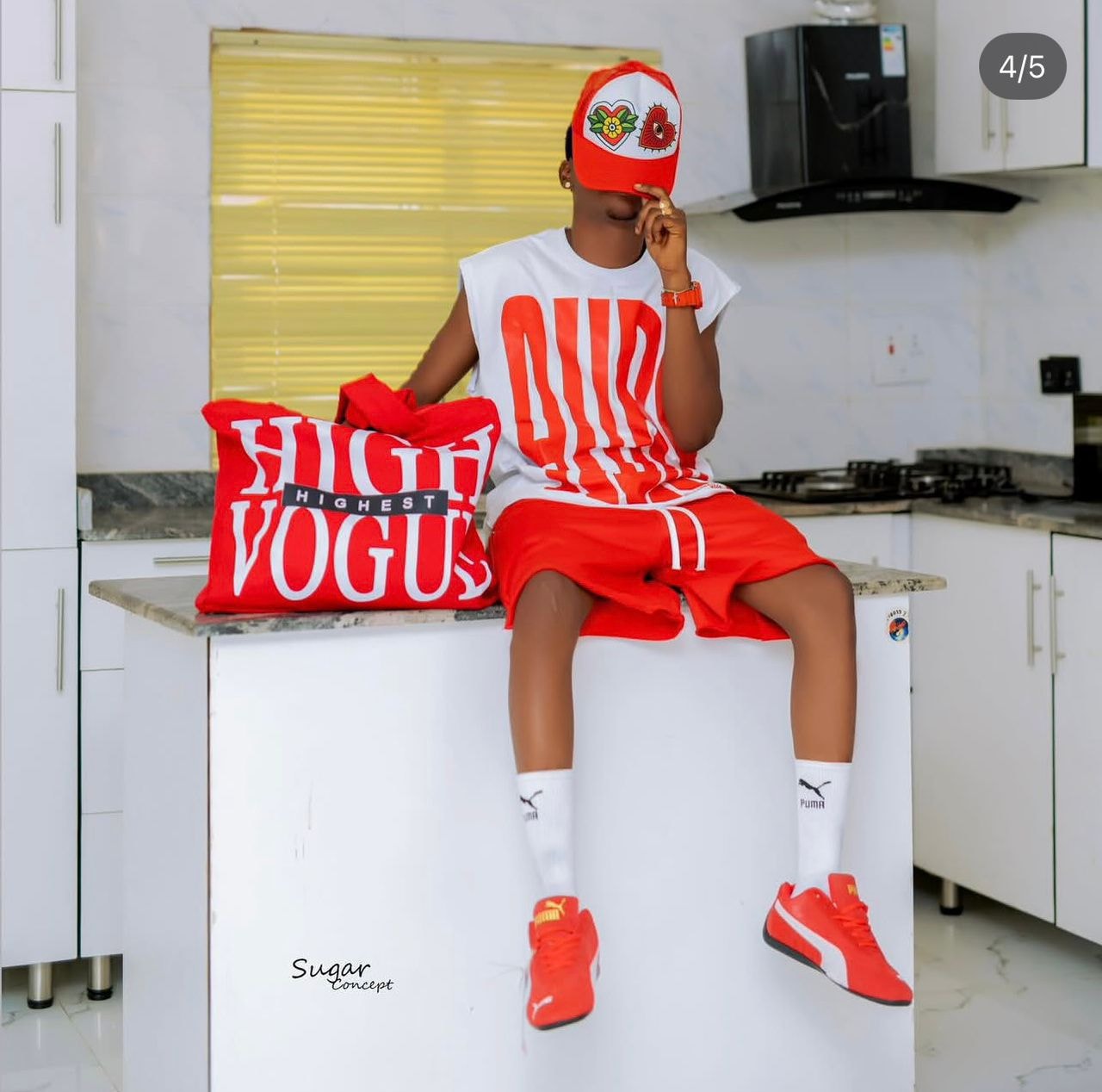 Model seated wearing white sleeveless top with red graphic print, red shorts, red cap and holding red HIGH VOGUE bag.