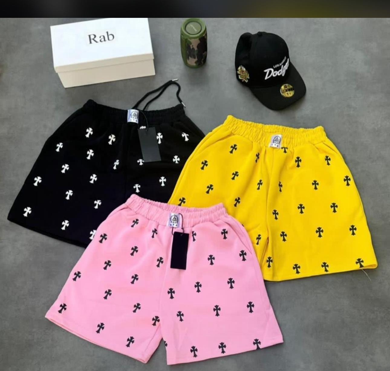 Pink, yellow and black cross print shorts arranged with cap and accessories on gray surface.