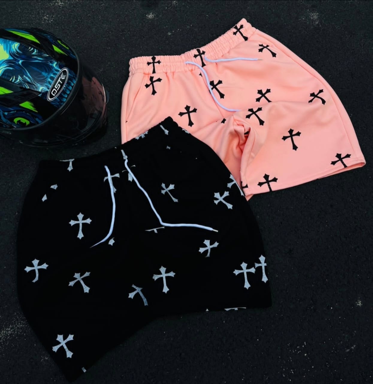 Black and pink cross print shorts displayed on asphalt next to a helmet.
