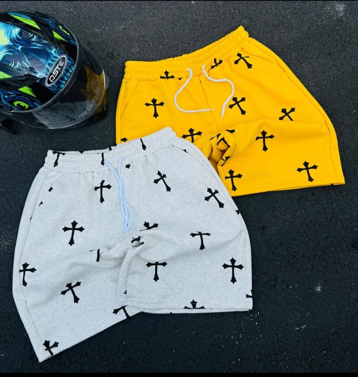 Yellow and gray cross print shorts laid on dark pavement beside a helmet.