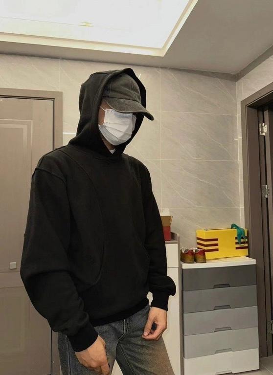 Man wearing black oversized hoodie with cap and face mask in modern indoor setting.