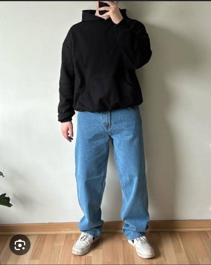 Man in black oversized hoodie paired with blue jeans and white sneakers, standing against a wall.