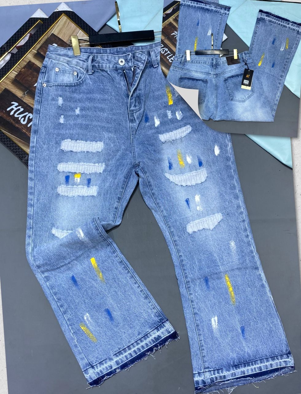 Blue distressed baggy jeans with rips, white patches, yellow and blue details shown in flat lay.