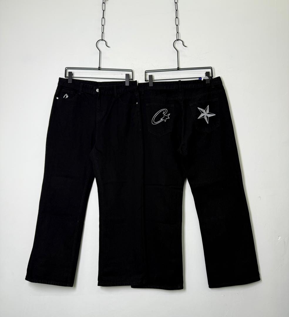 Two pairs of black jeans hanging, featuring white star and script embroidery on back pockets.