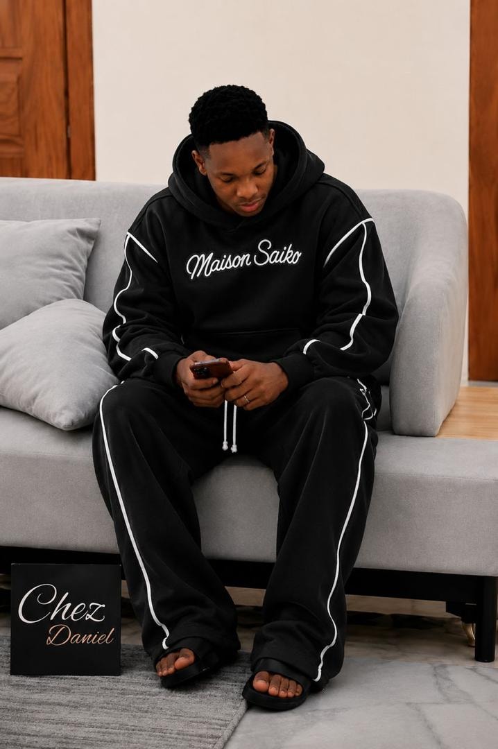 Man seated on couch wearing black oversized Maison Saiko hoodie and pants with white contrast piping.
