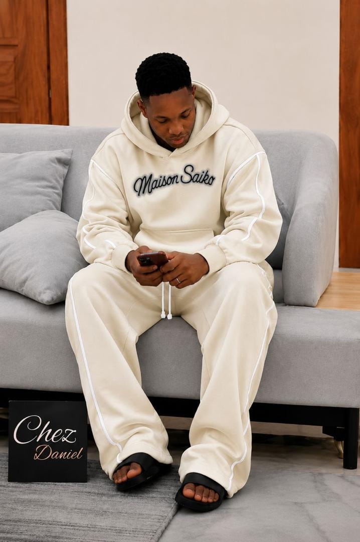 Man seated on couch wearing cream oversized Maison Saiko hoodie and pants with white contrast piping.