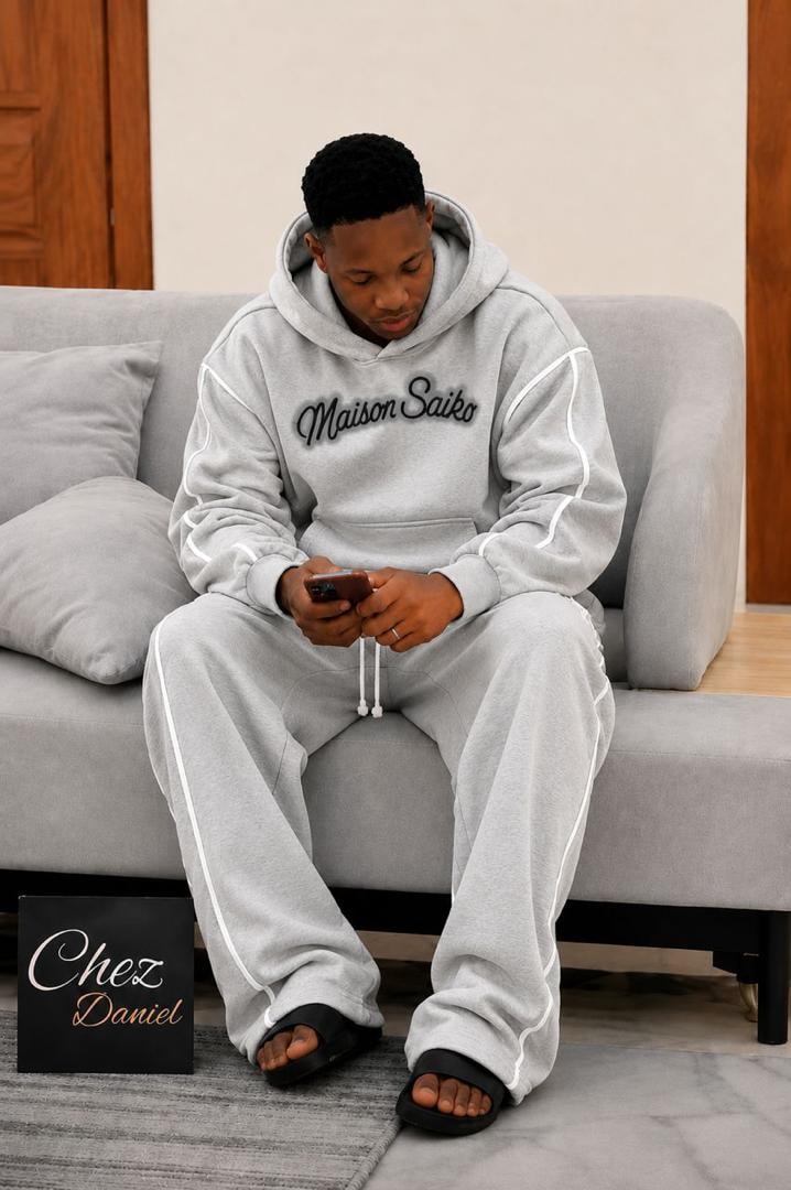 Man seated on couch wearing gray oversized Maison Saiko hoodie and pants with white contrast piping.