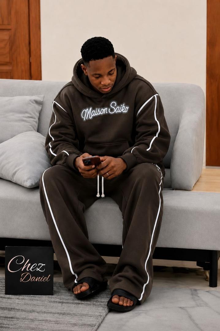 Man seated on couch wearing brown oversized Maison Saiko hoodie and pants with white contrast piping.