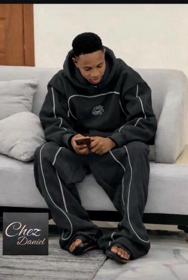 Man seated on couch wearing black oversized hoodie and pants with white contrast piping and circular chest emblem.
