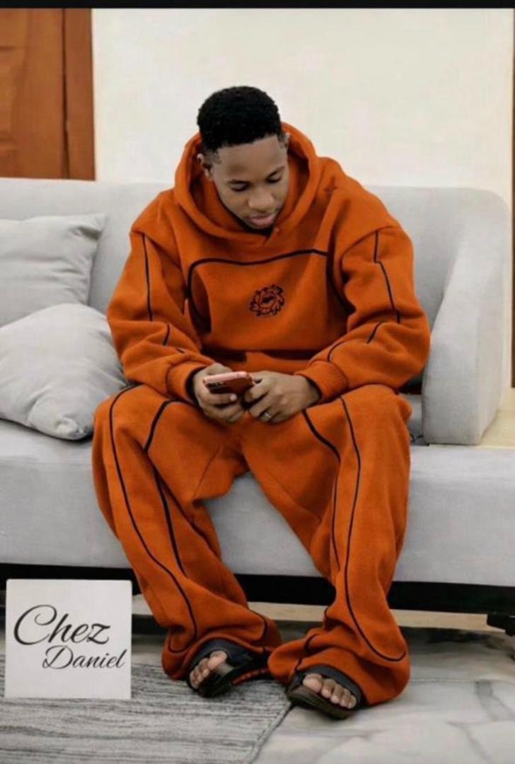 Man seated on couch wearing orange oversized hoodie and pants with black contrast piping and circular chest emblem.