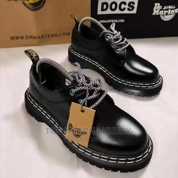 Pair of black Dr. Martens shoes with white stitching and branded box in background.