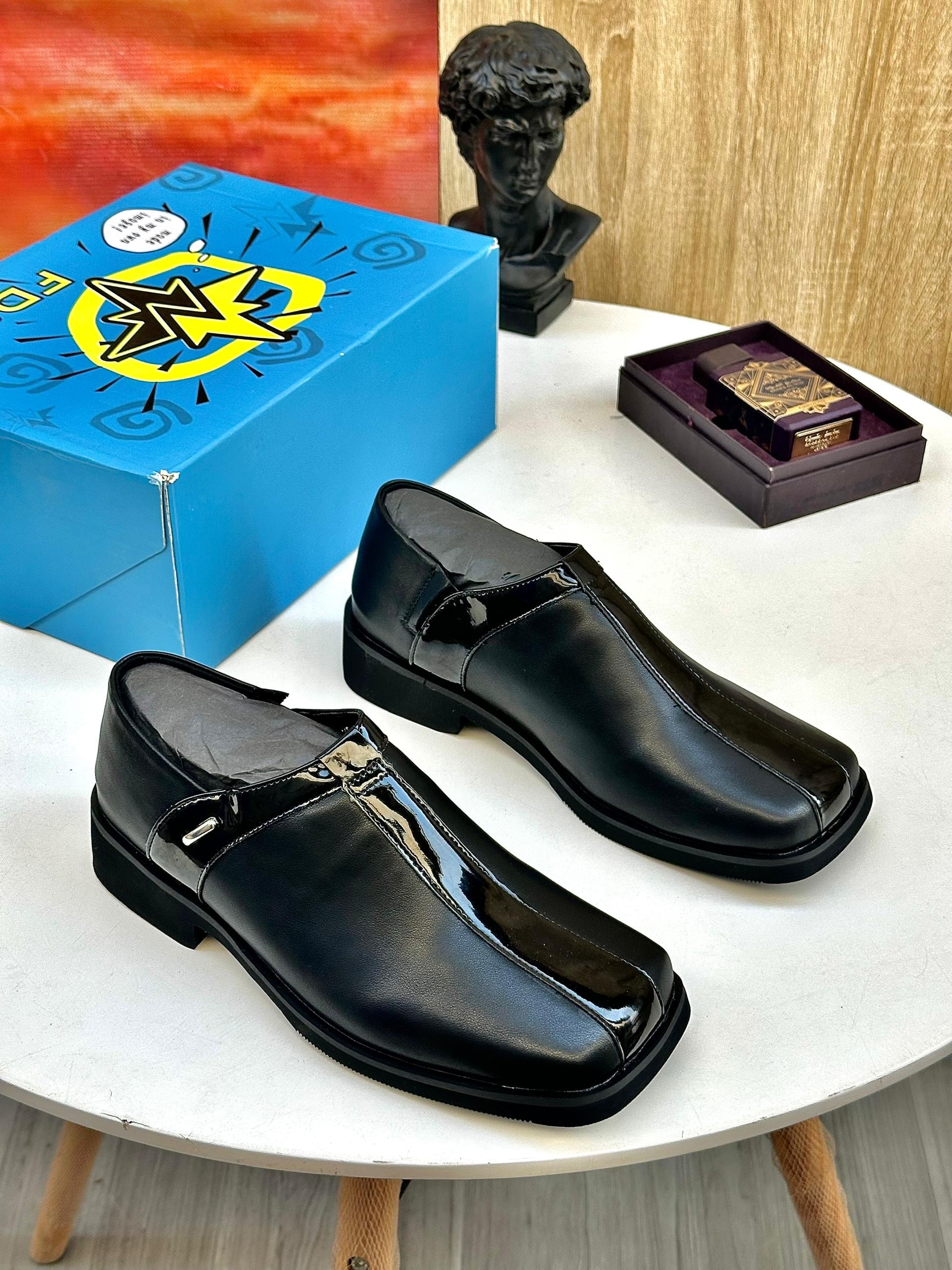 Pair of black patent leather slip-on loafers displayed on white table with blue box and bust sculpture.